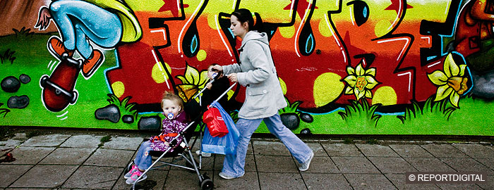 Young white woman walking with pushchair past graffiti wall