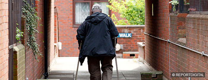Elderly man, walking with crutches, along walkway in flats, view from rear